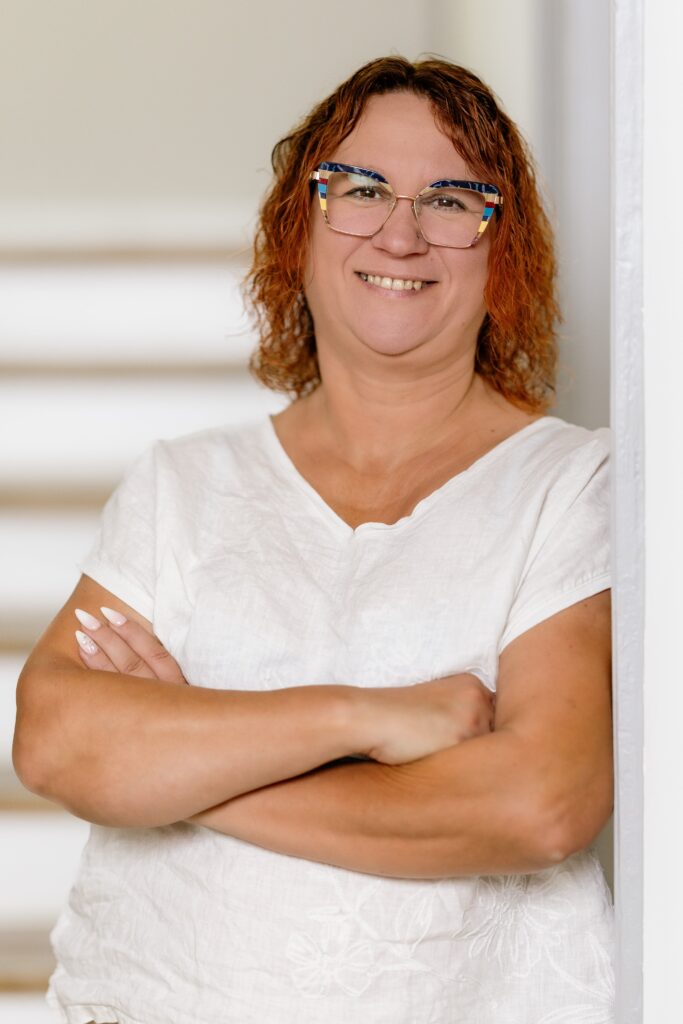 Lächelnde Frau mit lockigem, dunkelblondem Haar und farbigen Brillen, die in einem hellen Raum steht.