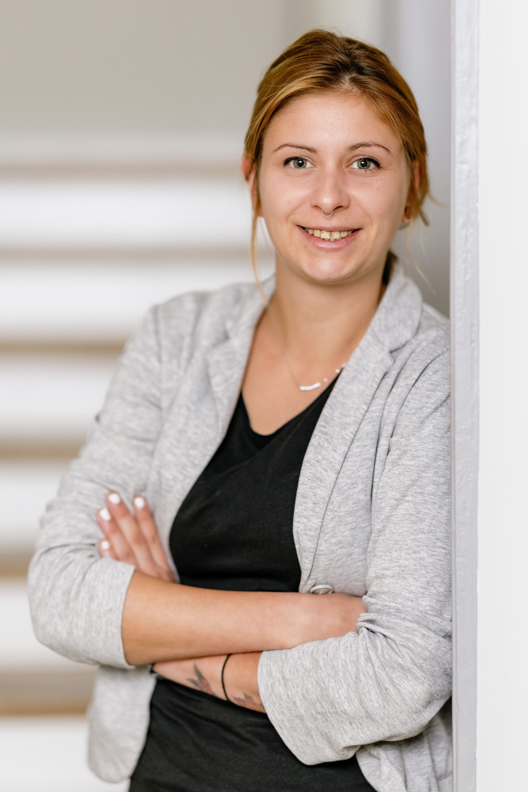 Lächelnde Frau mit blonden Haaren, gekleidet in graue Jacke, lehnt an einer Wand und hat die Arme verschränkt.