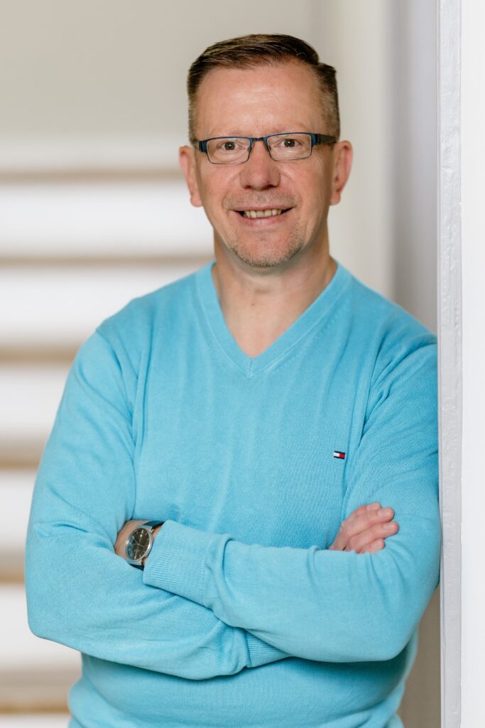 Ein lächelnder Mann in einem hellblauen Pullover mit verschränkten Armen steht vor einer hellen Wand.