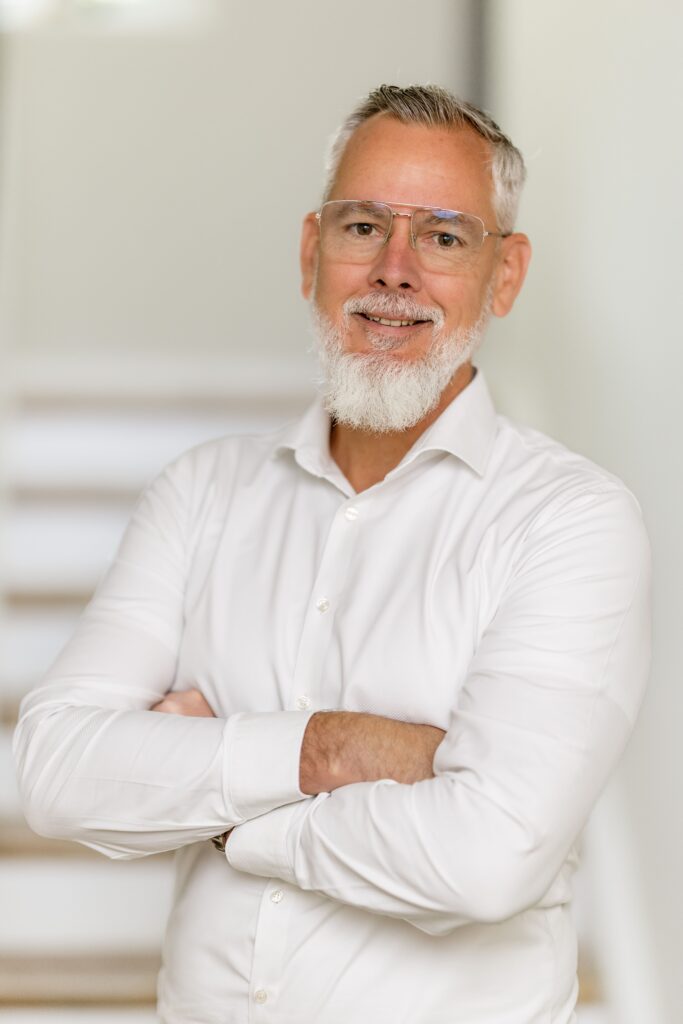 Mann mit Brille und Bart in einem weißen Hemd steht mit verschränkten Armen vor einer Treppe.