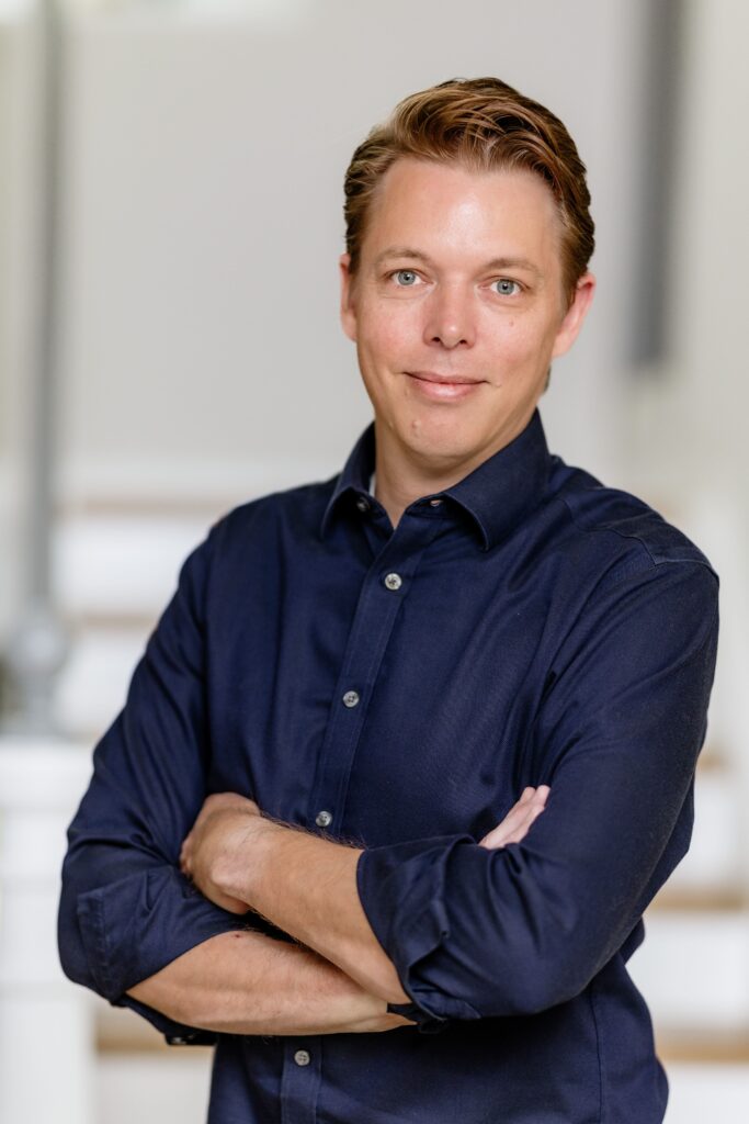 Ein Mann mit kurzgeschnittenen Haaren steht mit verschränkten Armen und lächelt in die Kamera. Er trägt ein dunkelblaue...