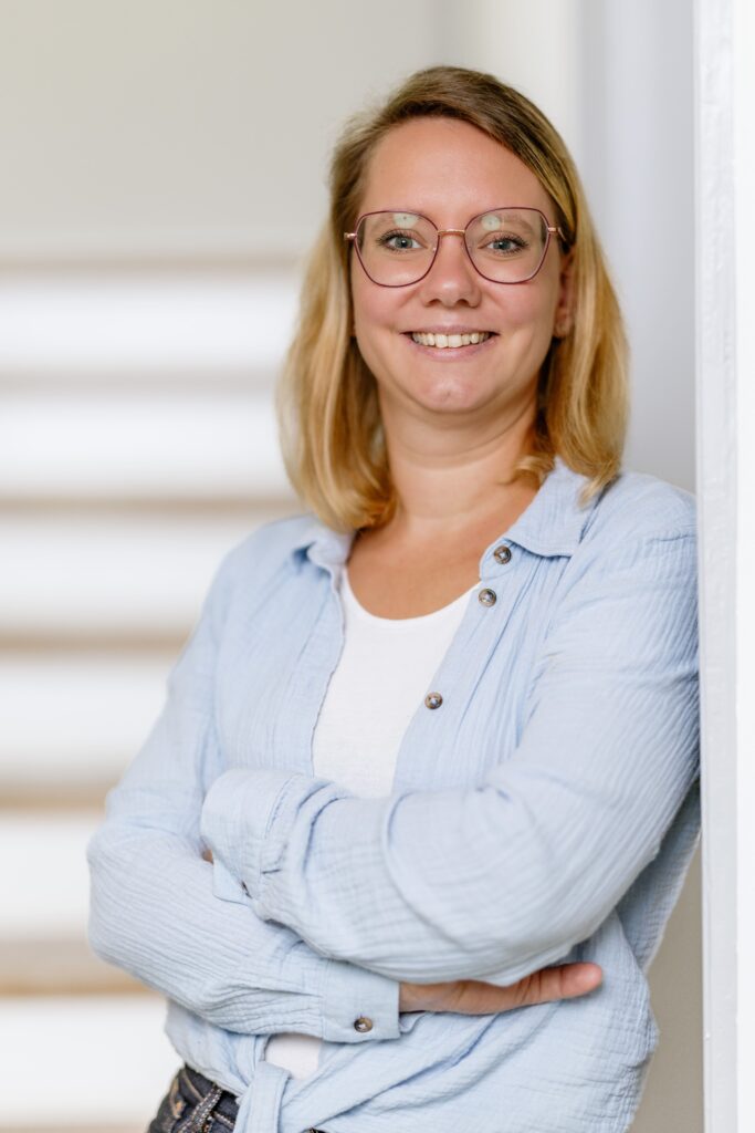 Lächelnde Frau mit blonden Haaren und Brille in einem hellen Raum, die entspannt an einer Wand steht.